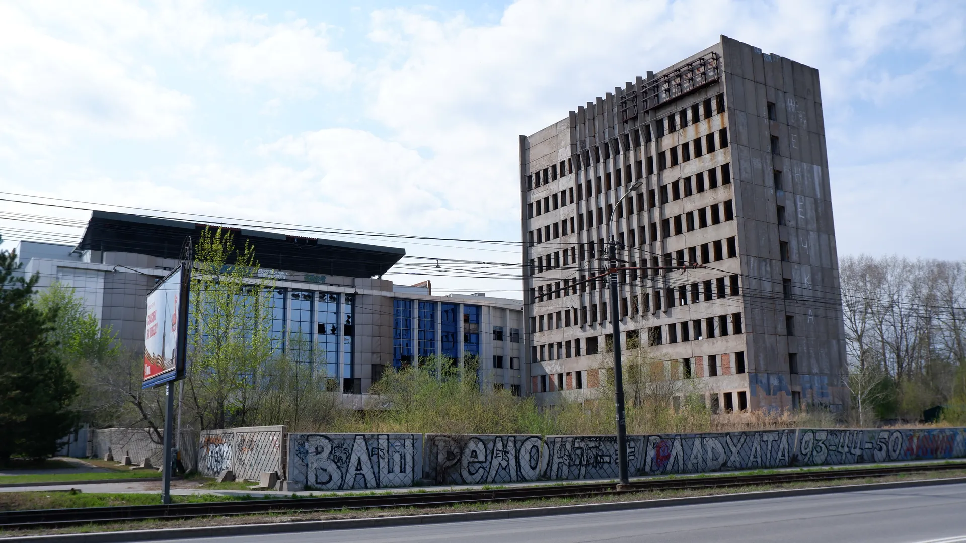 An abandoned high-rise building with a worn-out exterior and missing windows. In the foreground, there is a graffiti-covered wall and a railway track. In the background, there are trees and another modern building.