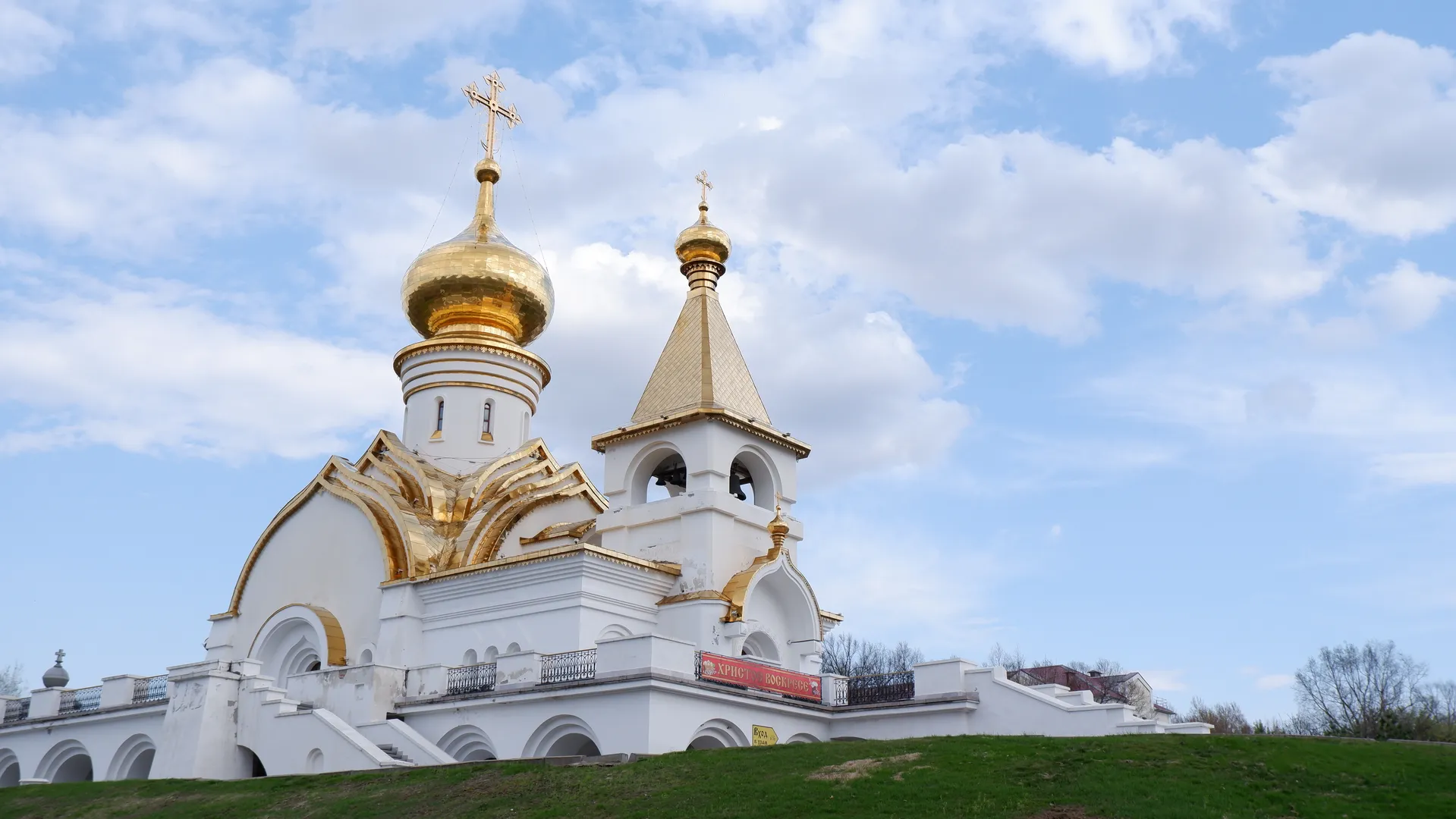 A white church with a golden dome sits on a meadow. The church has a cross on top, and there are red banners on its walls. The sky has white clouds.