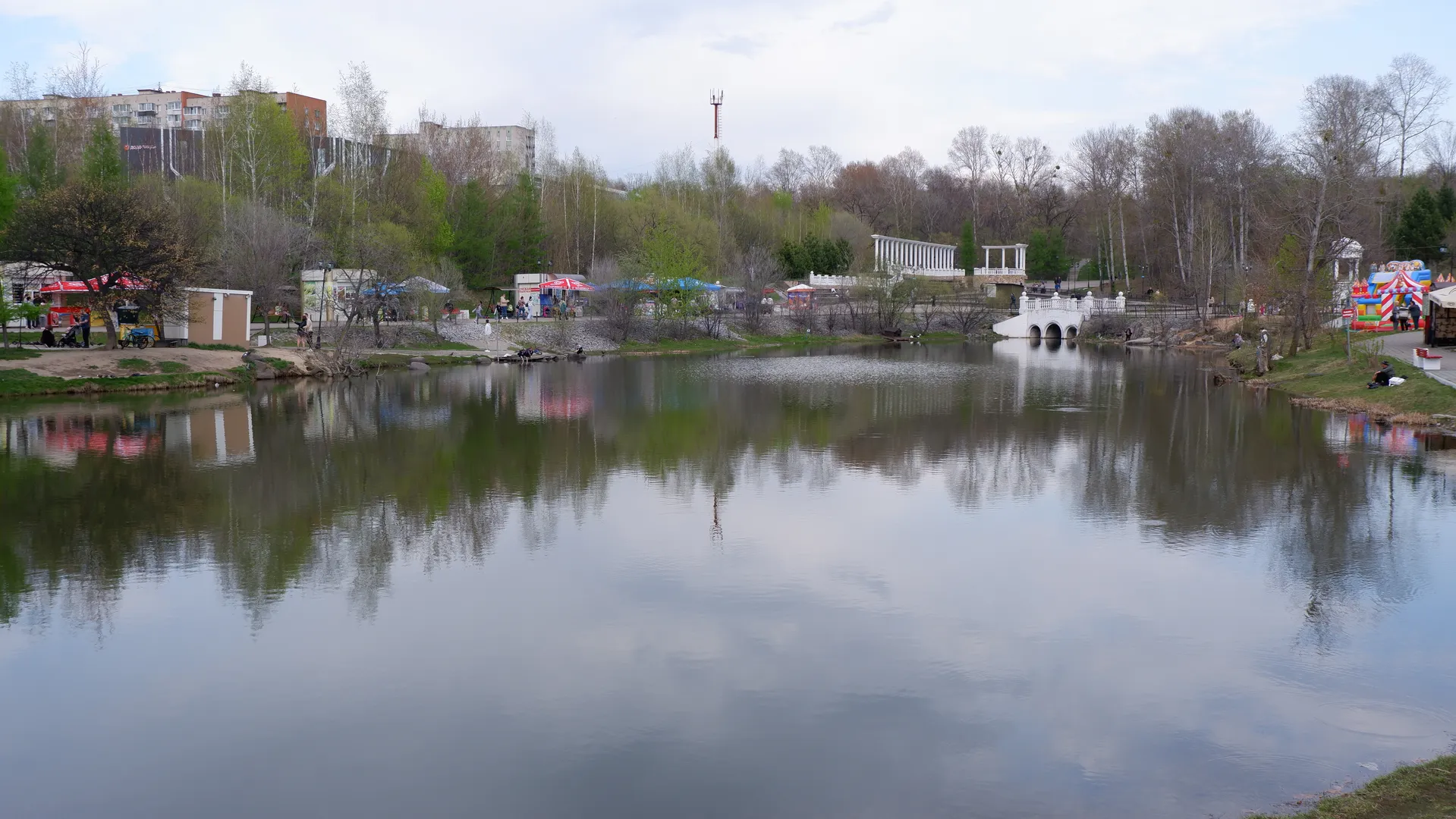 A tranquil lake reflects the surrounding scenery, with trees and buildings at the lakeside. In the distance, some amusement facilities and tents can be seen. The sky is overcast.