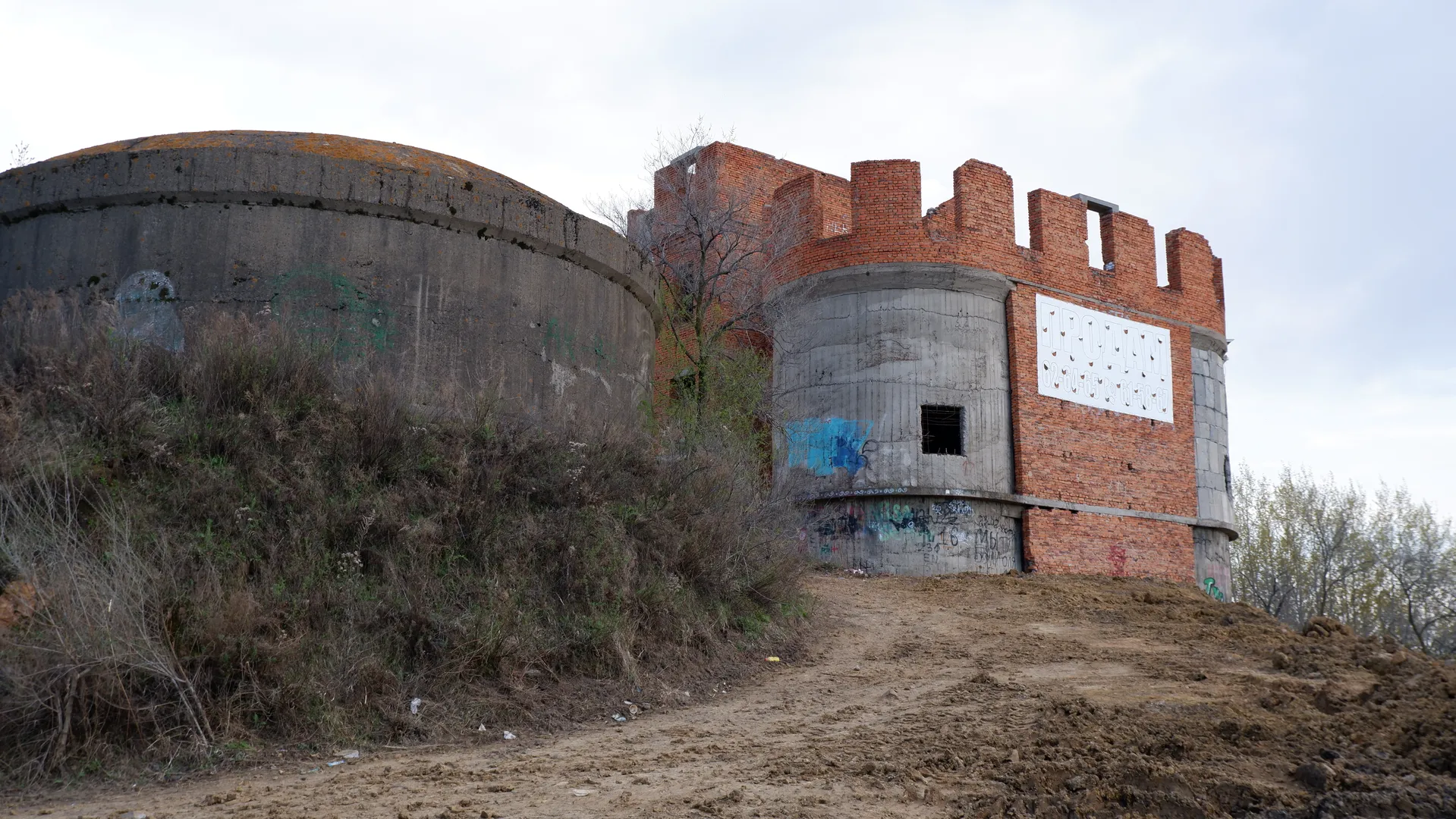 An unfinished brick building sits on a dirt slope, with some parts made of concrete. The building is covered with graffiti, and the surrounding area is overgrown with weeds. The sky is overcast.