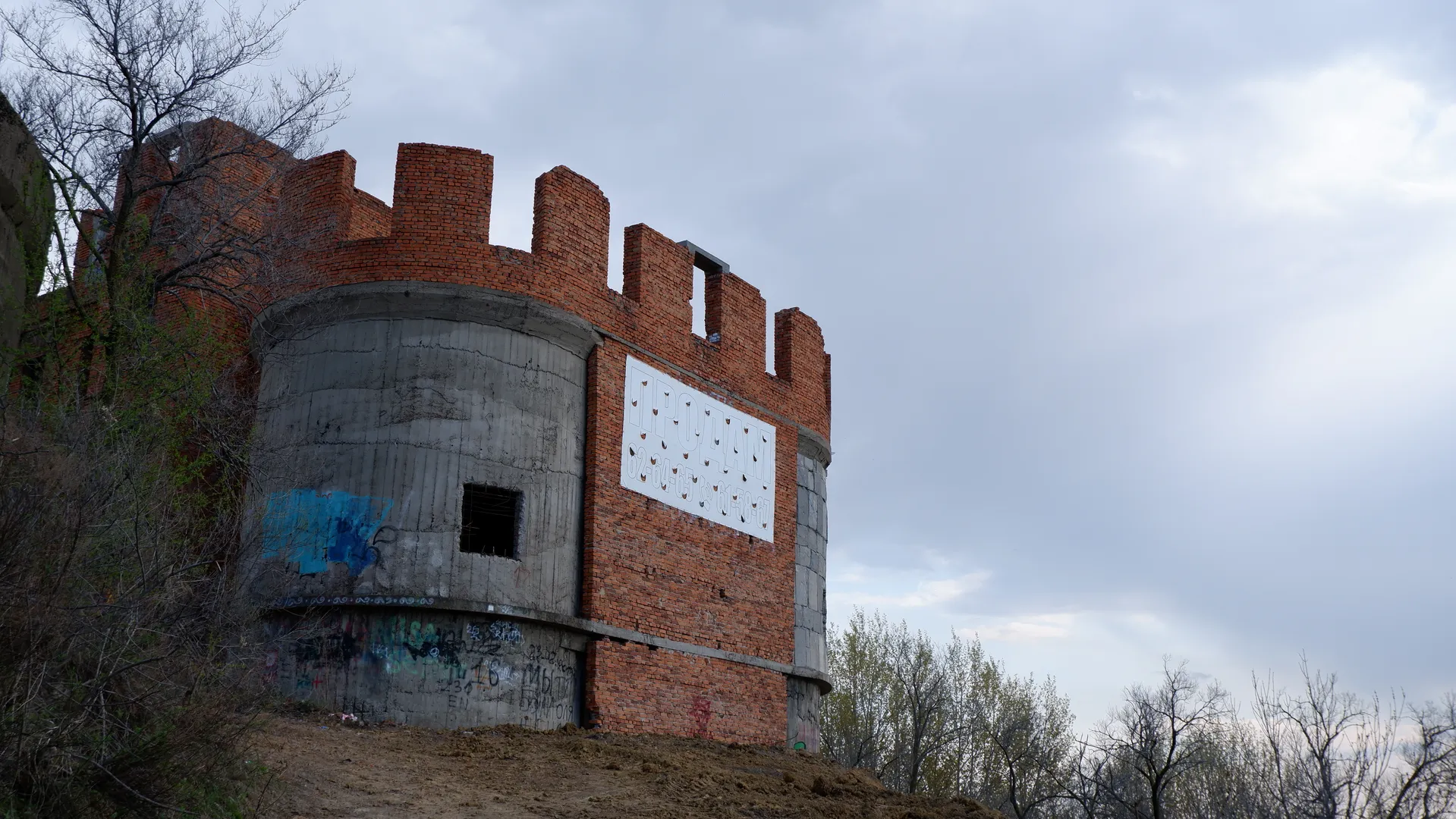 The tower of a red-brick building is visible, with square windows on the tower. The tower has graffiti and a white sign. In the background, there are trees and a cloudy sky.