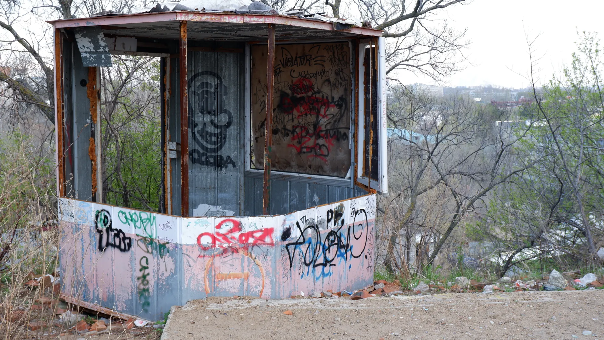 A small metal kiosk is covered in graffiti, with broken glass windows. The kiosk is surrounded by dirt and vegetation, and in the background, trees and distant buildings can be seen. The sky is overcast.