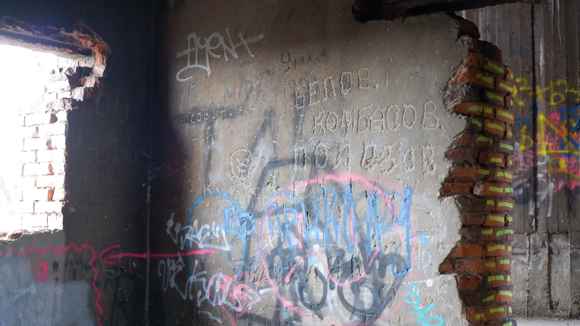 A wall covered in various graffiti, including blue and pink patterns and some Russian text. Part of the wall is made of bricks, showing significant wear. Sunlight streams from the left, illuminating part of the wall.