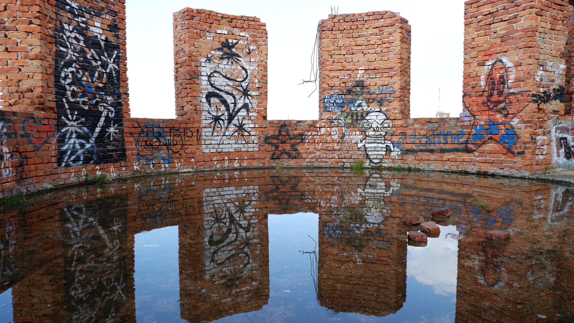 A red brick wall submerged in water, with colorful graffiti patterns. The structure of the wall is partially damaged, with several protruding columns. The water is calm, reflecting the graffiti on the wall.