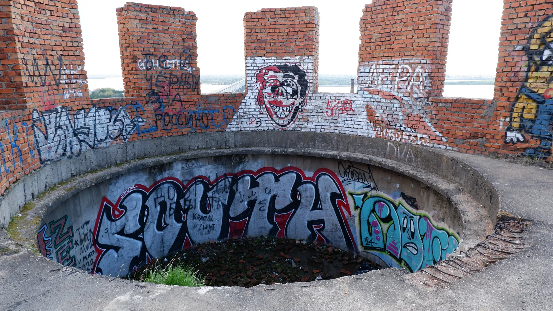 A circular recessed area surrounded by a red-brick wall, covered in vibrant graffiti. Inside the recess, there are more graffiti, mainly red and blue letters. In the background, distant rivers and the sky can be seen.