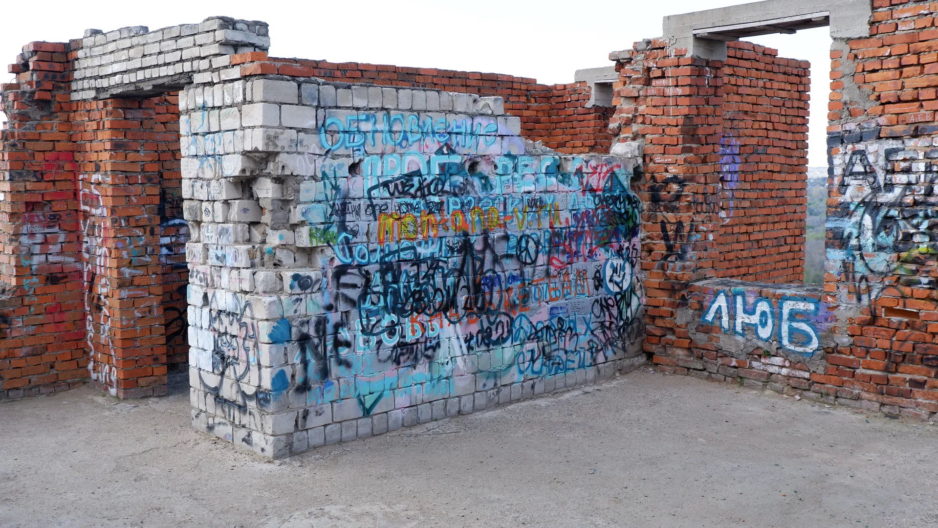 The wall of a ruin is covered with dense graffiti in various colors. The wall is made of a mix of red and white bricks, with parts of the wall collapsed. The ground is bare dirt, giving a desolate feel.