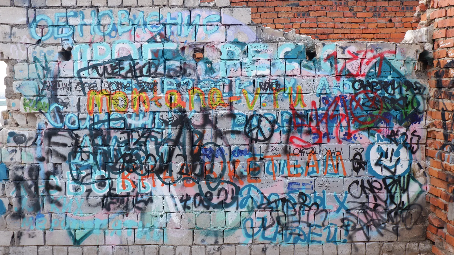 A white brick wall covered in colorful graffiti, with intricate patterns. The graffiti includes many letters and symbols, along with simple shapes. Some parts of the wall are damaged, exposing the texture of the bricks.