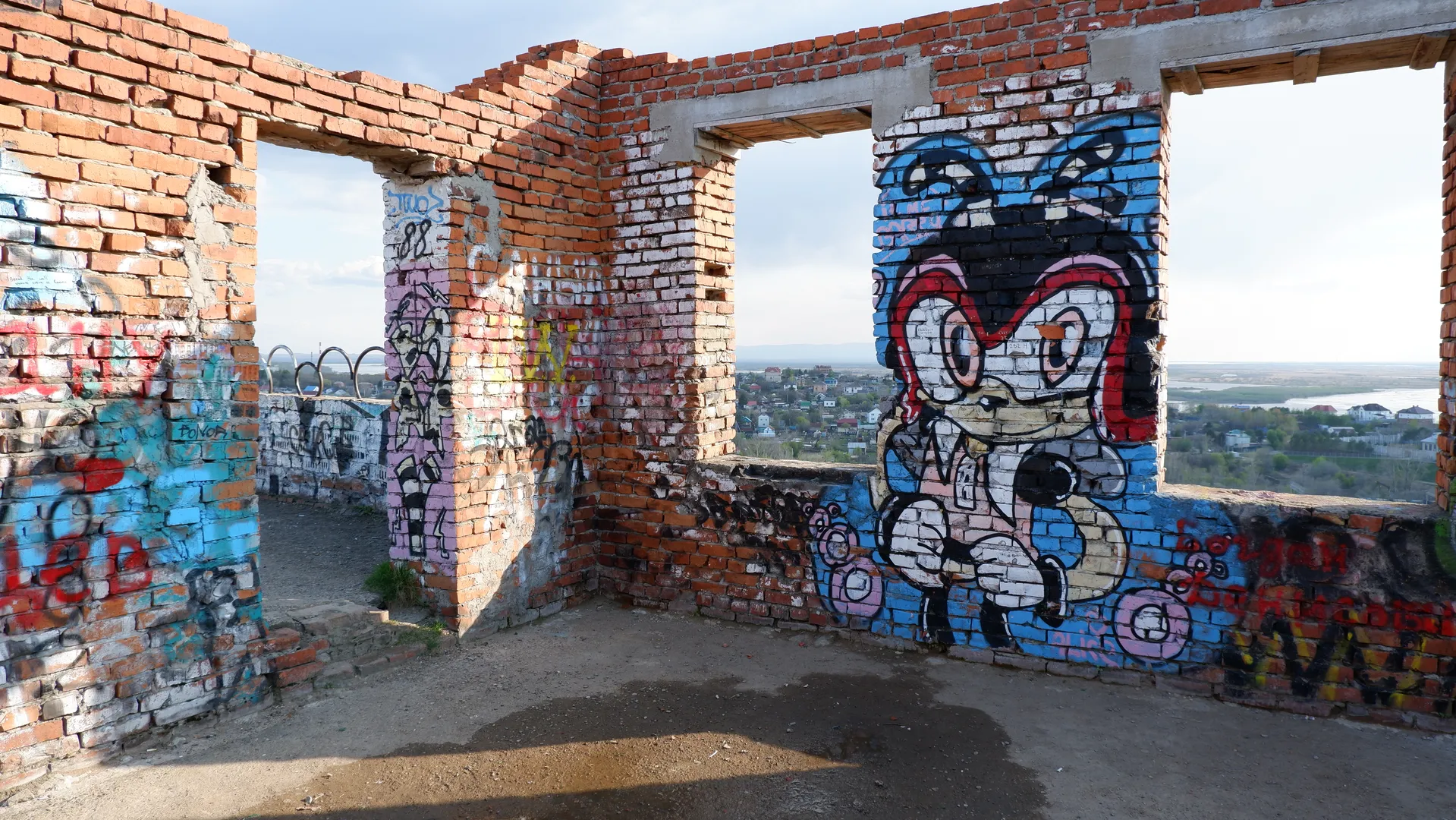 On the remains of an abandoned building, there is a large cartoon graffiti depicting a character with a red headband. The red brick walls and concrete structure of the building are visible. Through the gap, distant cityscapes and rivers can be seen.