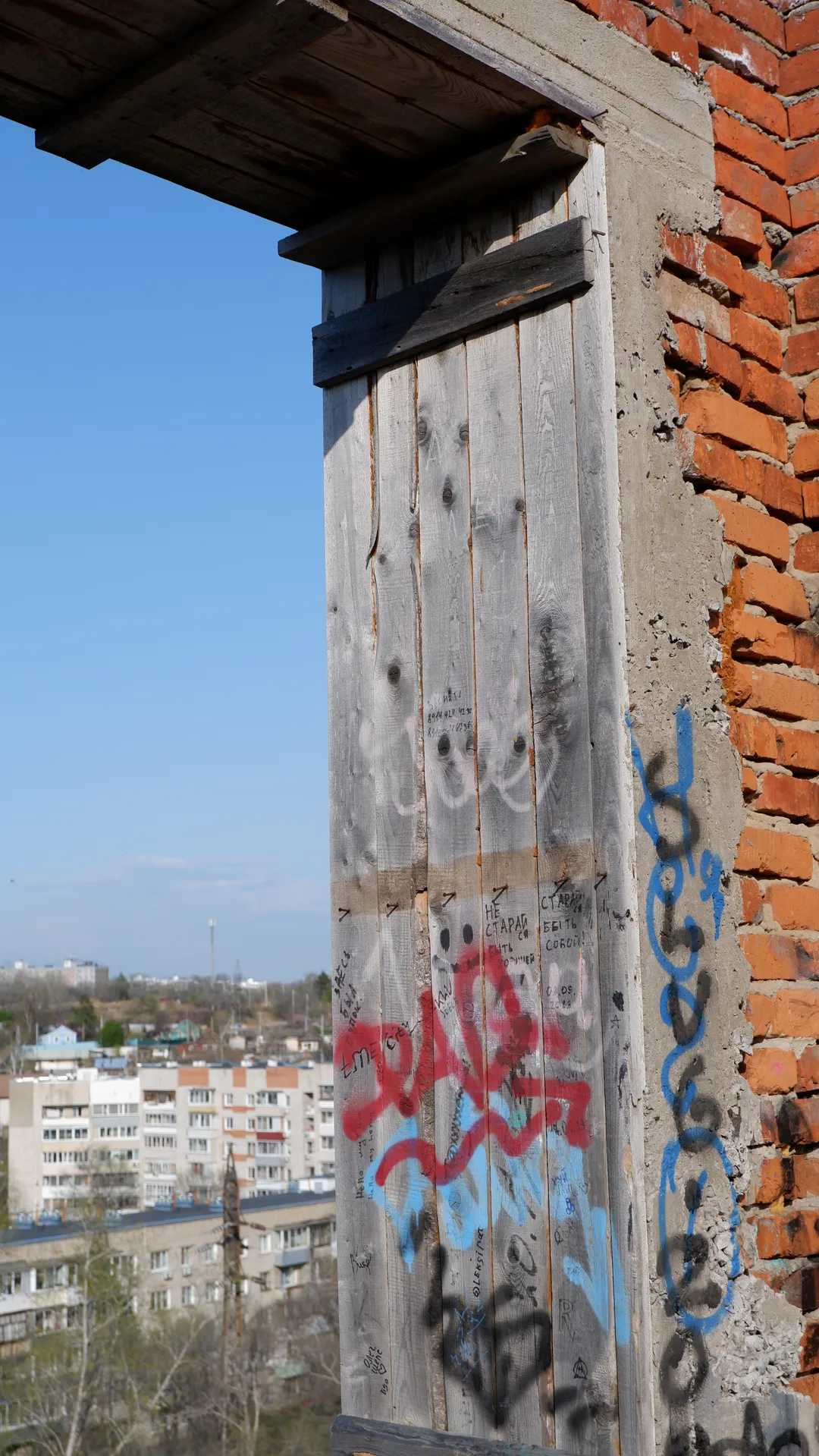 A wooden window with some graffiti on it, made from several vertical wooden planks. Next to the window is a red brick wall, also with some graffiti. Outside the window, there are city buildings and blue skies.