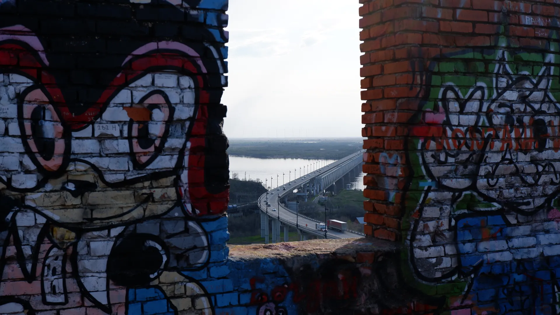 Between two graffiti-covered walls, a bridge can be seen, with vehicles passing over it. The graffiti on the walls is colorful, with one of the graffiti showing a large-eyed character. The bridge spans across water, connecting two shores.