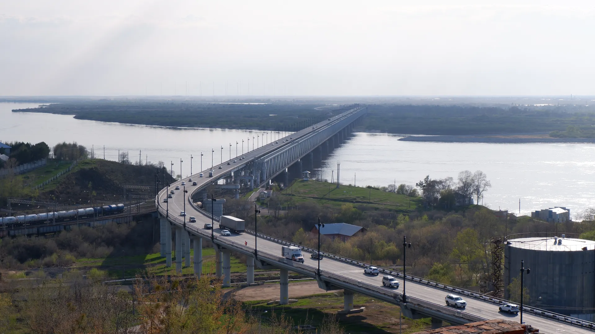 A long bridge spans over water, with multiple lanes for vehicles. Streetlights and guardrails line both sides of the bridge, and beneath the bridge, there is a wide river. In the distance, flat terrain and some vegetation can be seen.