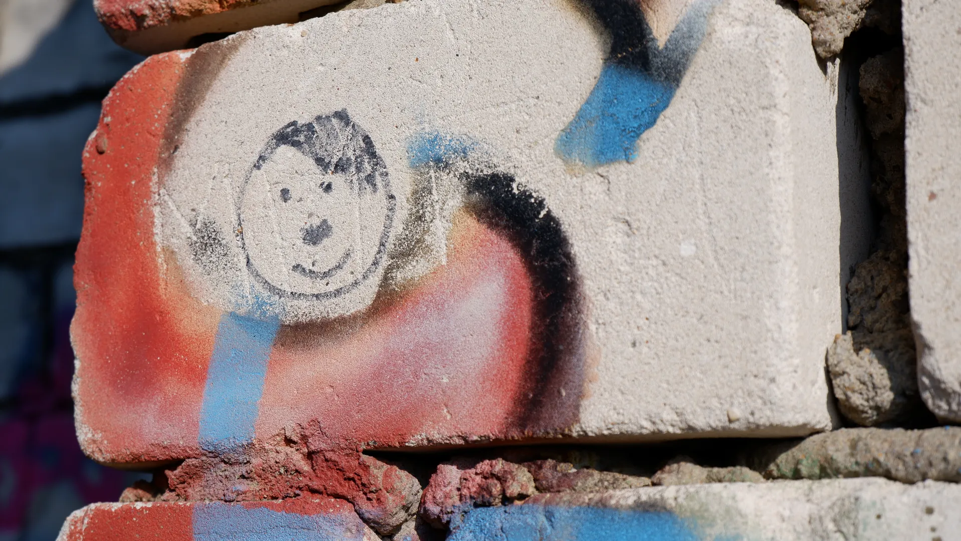 A brick with a simple smiley face graffiti drawn in black lines. The brick is mainly red and gray, with some wear on the surface. Other colorful graffiti marks can be seen around the brick.