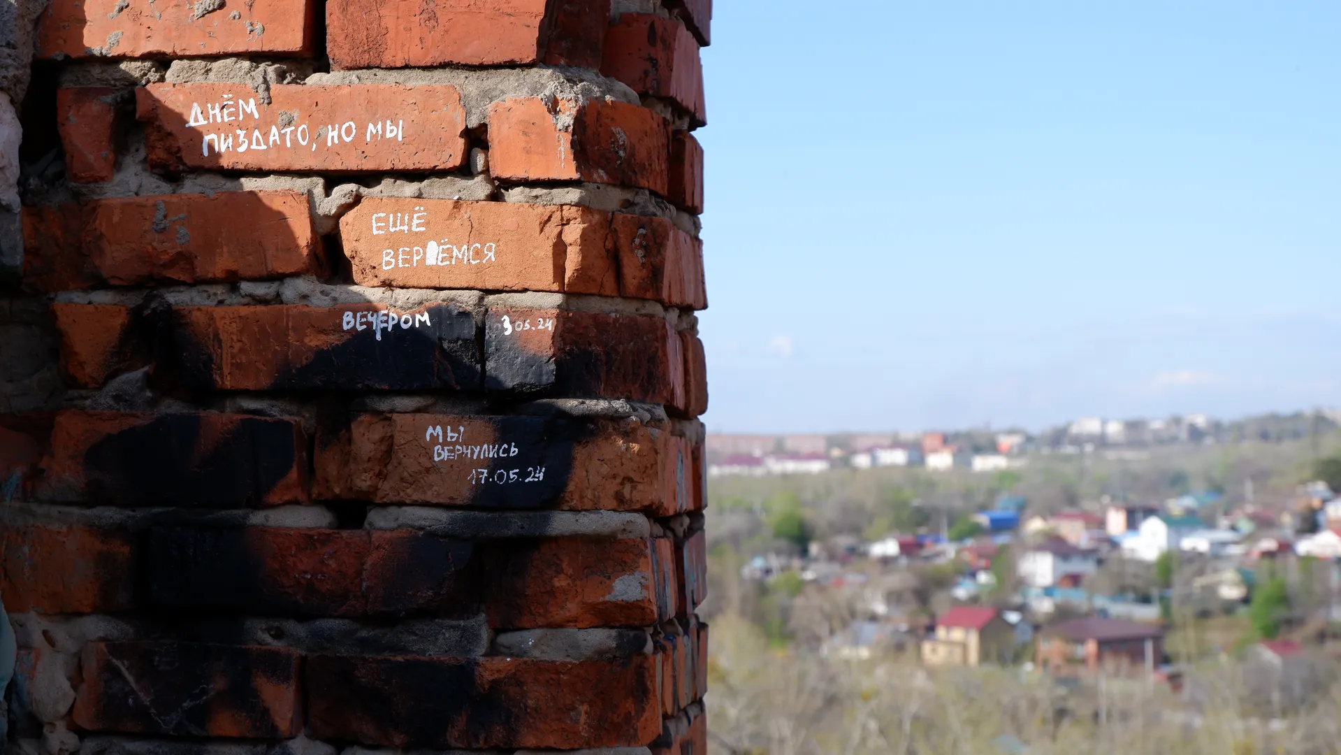 A brick wall with Russian graffiti, with distant houses and blue skies in the background. The text on the wall reads: “ДНЁМ ПИЗДАТО, НО МЫ ЕЩЁ ВЕРНЁМСЯ”. Some of the bricks are burnt black.