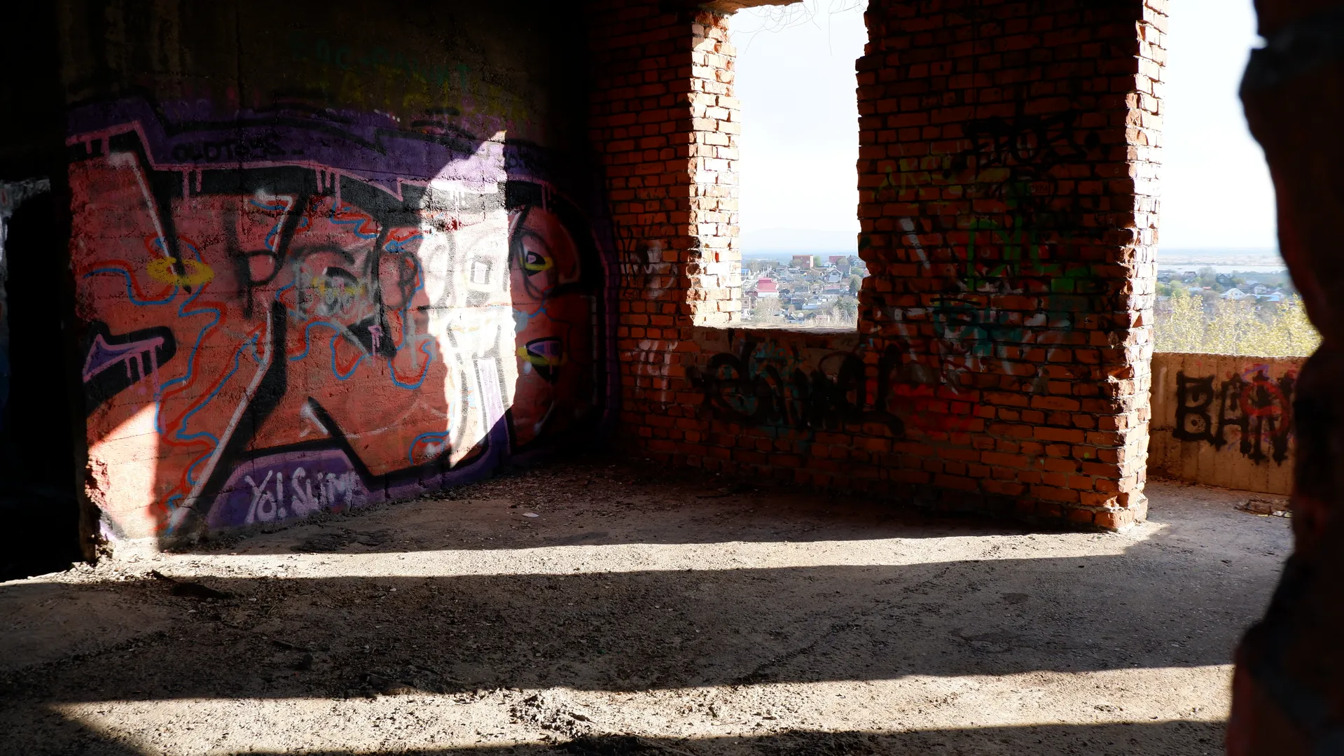 Inside an abandoned building, the walls are covered with graffiti. Through the window, the view outside can be seen. The floor is scattered with sandbags and debris. Sunlight streams in from the window.