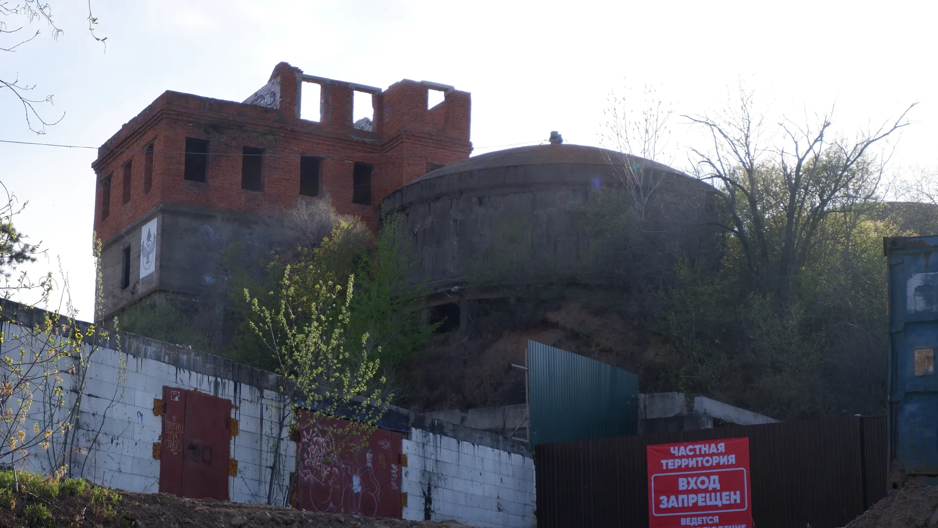A red brick building with partial damage, surrounded by trees and a fence. In front of the building is a red sign reading “ЧАСТНАЯ ТЕРРИТОРИЯ ВХОД ЗАПРЕЩЕН”. The building appears dilapidated.