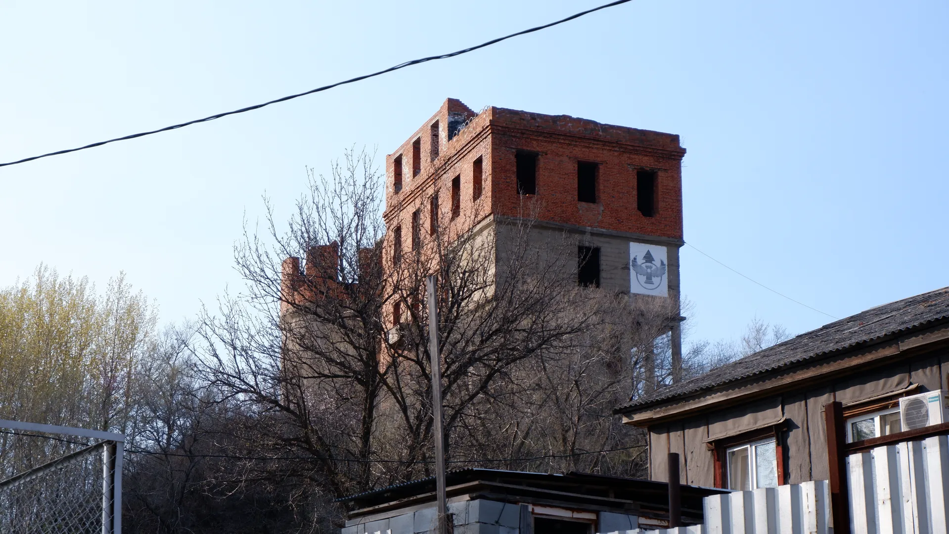 A tall red brick building with a damaged top, surrounded by bare trees. On the side of the building is a white sign with a pattern. The sky is clear.