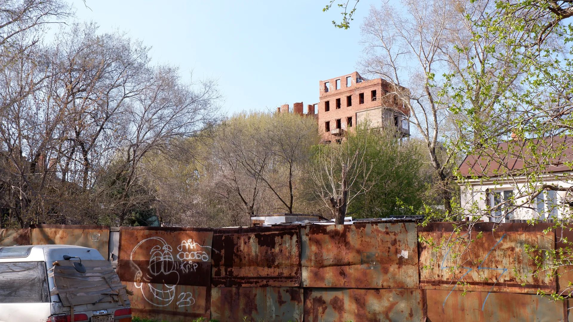 An abandoned building partially hidden behind trees, with a rusted fence in the foreground. The fence has graffiti, including a smiley face. Some of the trees are budding.