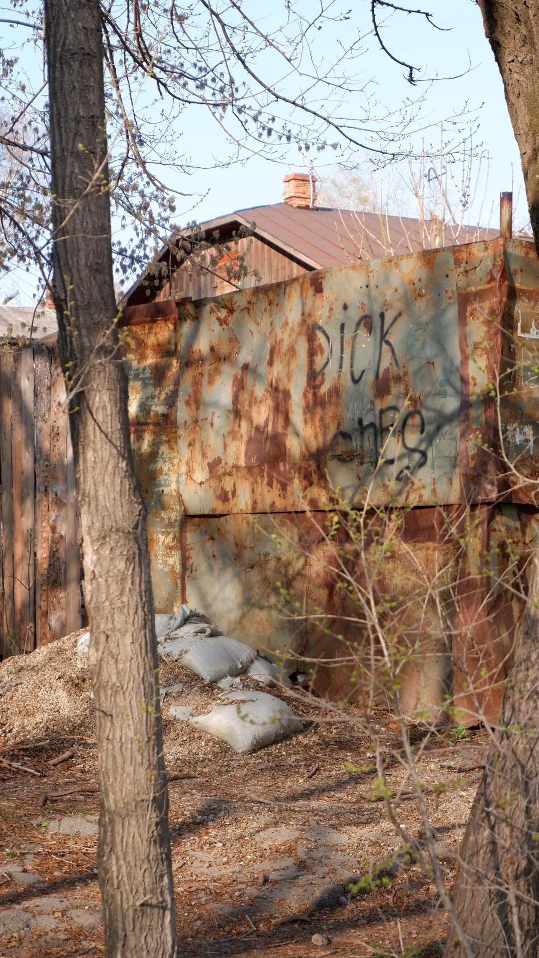 In front of a rusted wall are several trees. The wall has graffiti that reads “DICK”. Sandbags are piled up at the base of the wall, and the ground is cluttered.