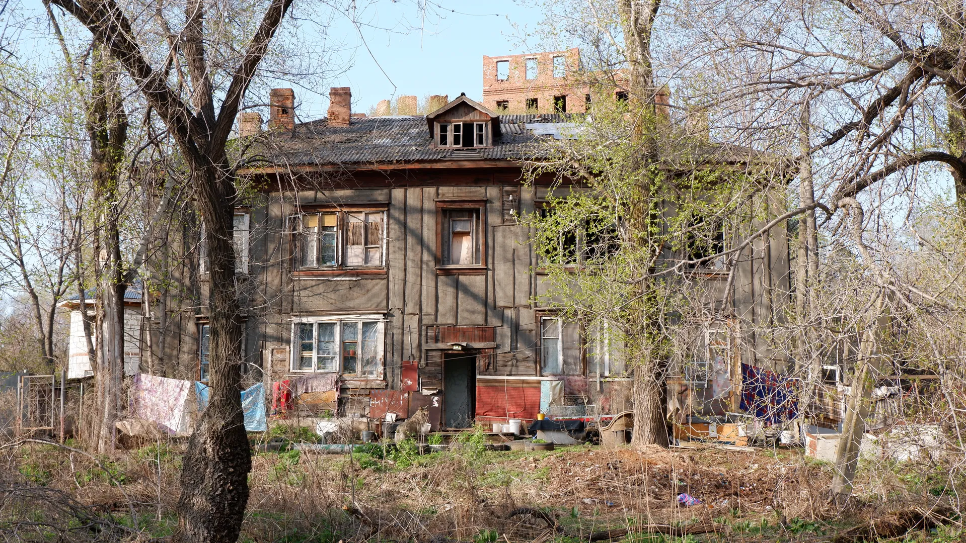 A two-story old residential building with a worn-out exterior and damaged windows. Weeds grow in front of the building, with some clothes hanging. In the background, there are more trees and an abandoned building.