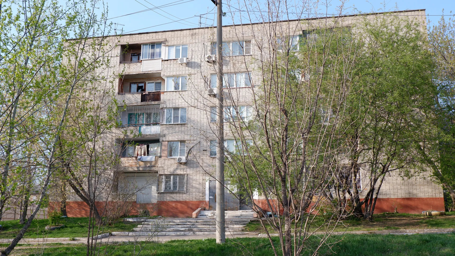 A multi-story residential building with light-colored brick exterior and neatly arranged windows. There are several trees in front of the building, with lush foliage, and stairs leading to the entrance. Power poles and wires cross the frame.
