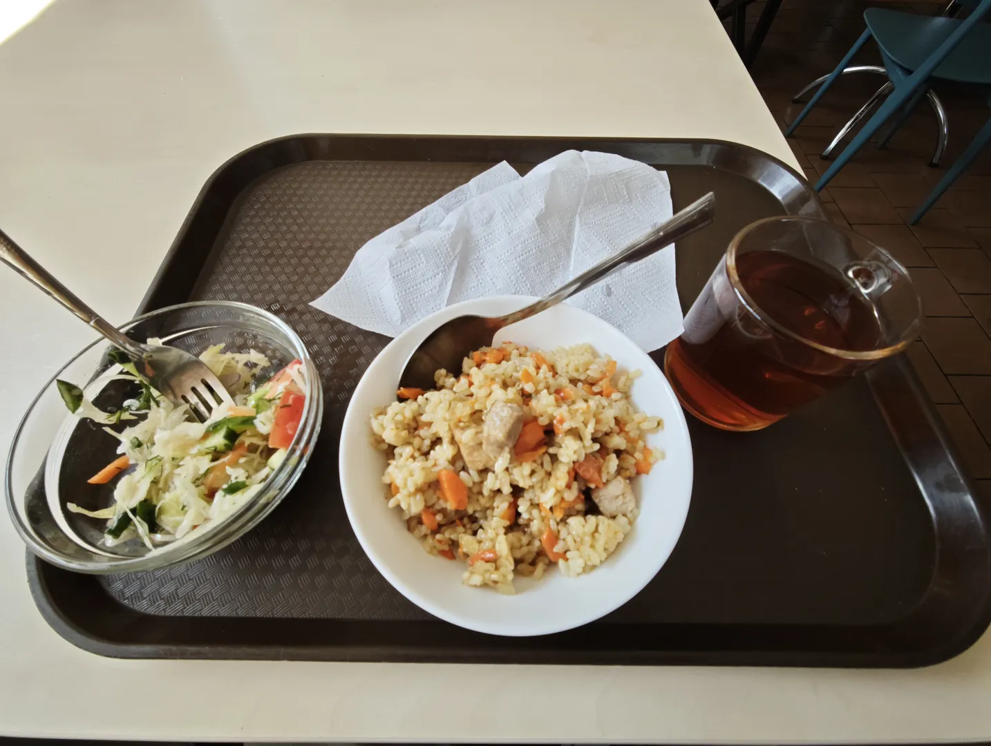 A tray with a bowl of rice, a bowl of salad, and a cup of tea. Next to it are napkins and utensils. The rice contains carrots and meat chunks, and the salad is made up of vegetables. The tray is placed on a table.