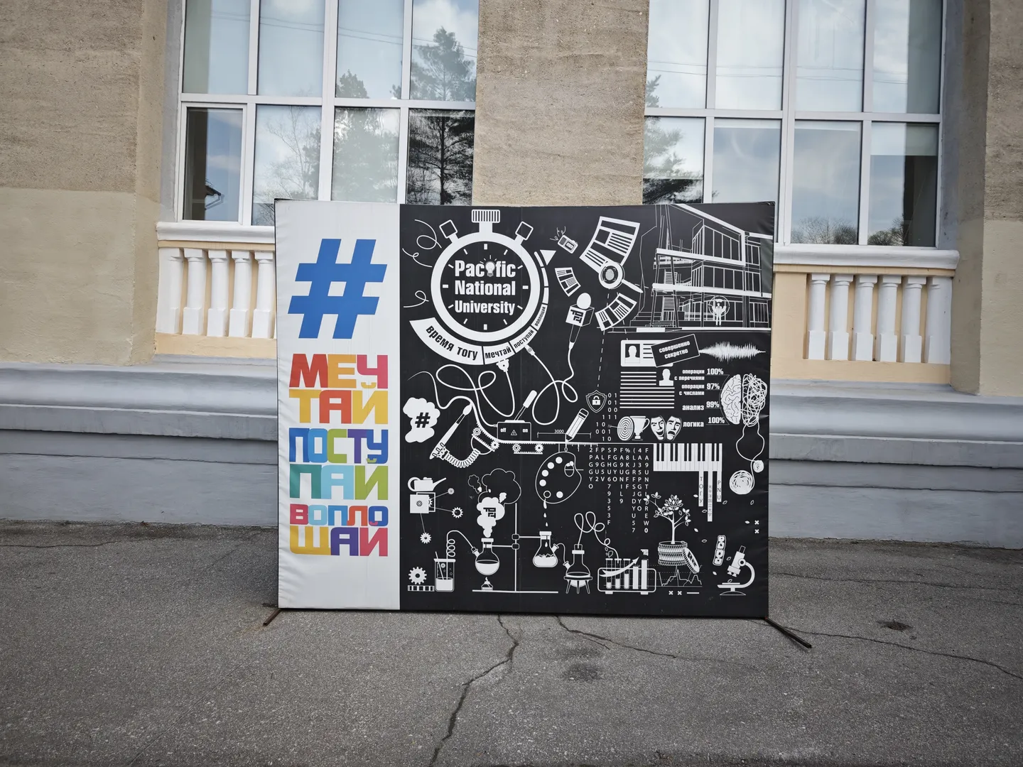 A billboard stands in front of a building. On the left side, there’s colorful text “МЕЧТАЙ ПОСТУПАЙ ВОПЛОЩАЙ”, and on the right, there are patterns and the words “Pacific National University”. The background of the billboard has a black-and-white pattern, and the building features large windows.