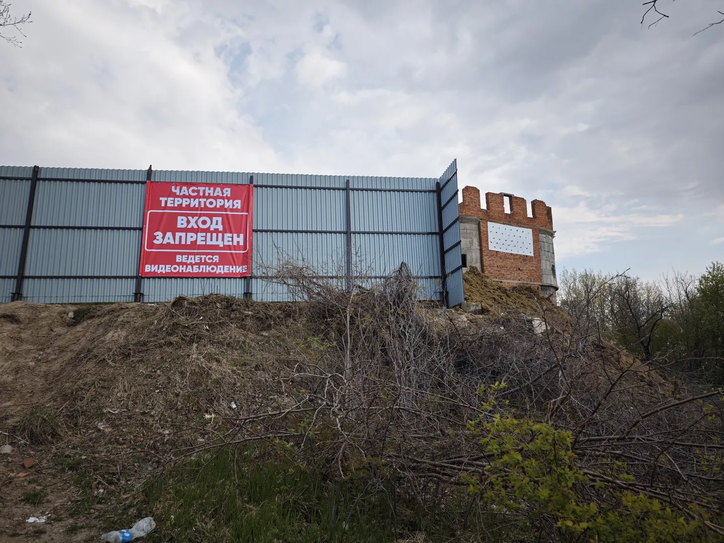 Behind a high wall stands a brick building. A red sign on the wall reads “ЧАСТНАЯ ТЕРРИТОРИЯ ВХОД ЗАПРЕЩЕН ВЕДЕТСЯ ВИДЕОНАБЛЮДЕНИЕ”. In front of the wall are weeds and dirt piles.