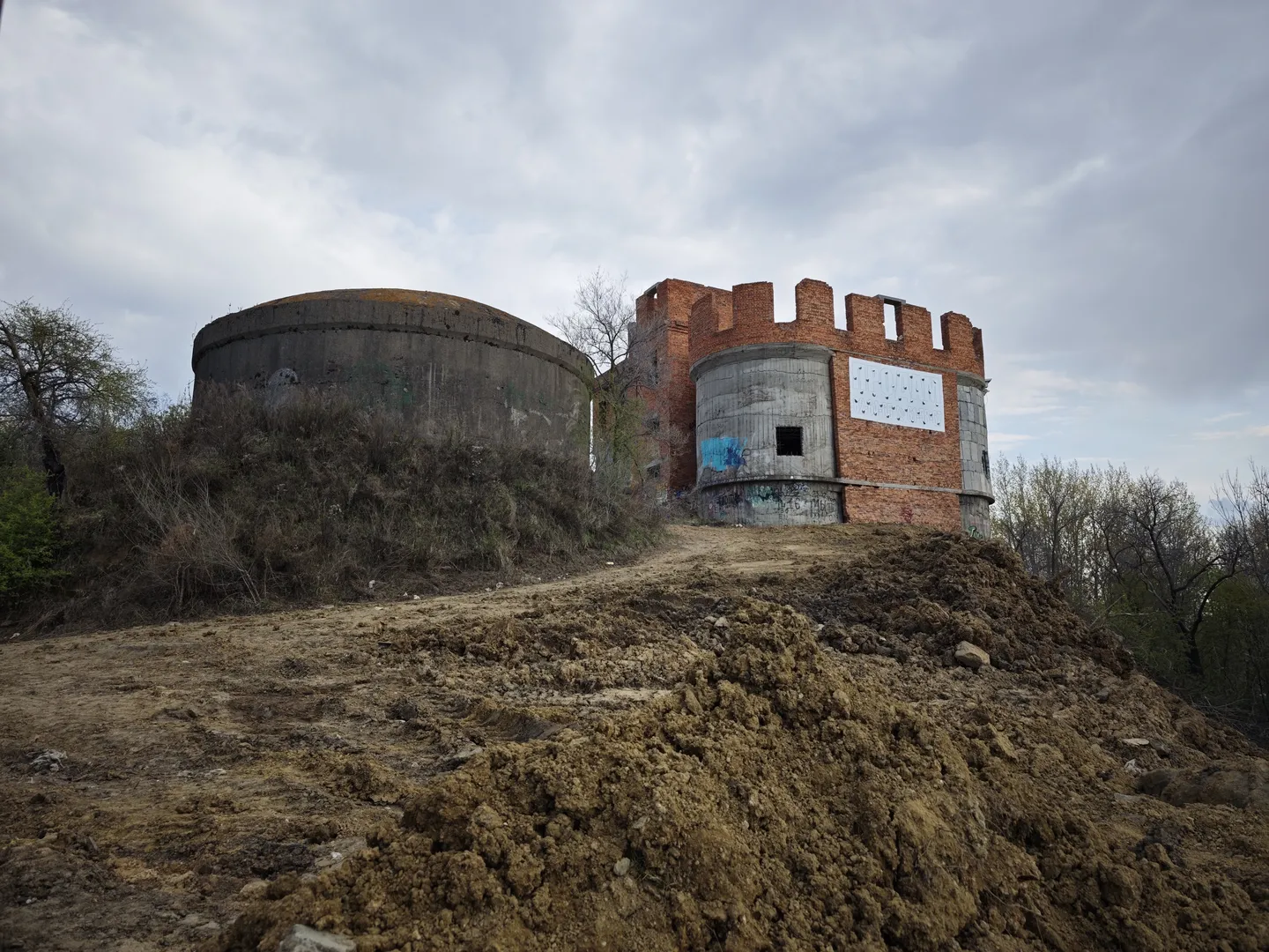An abandoned brick building on a dirt slope, partially damaged. The structure includes a dome and a tower, with graffiti on the surface.