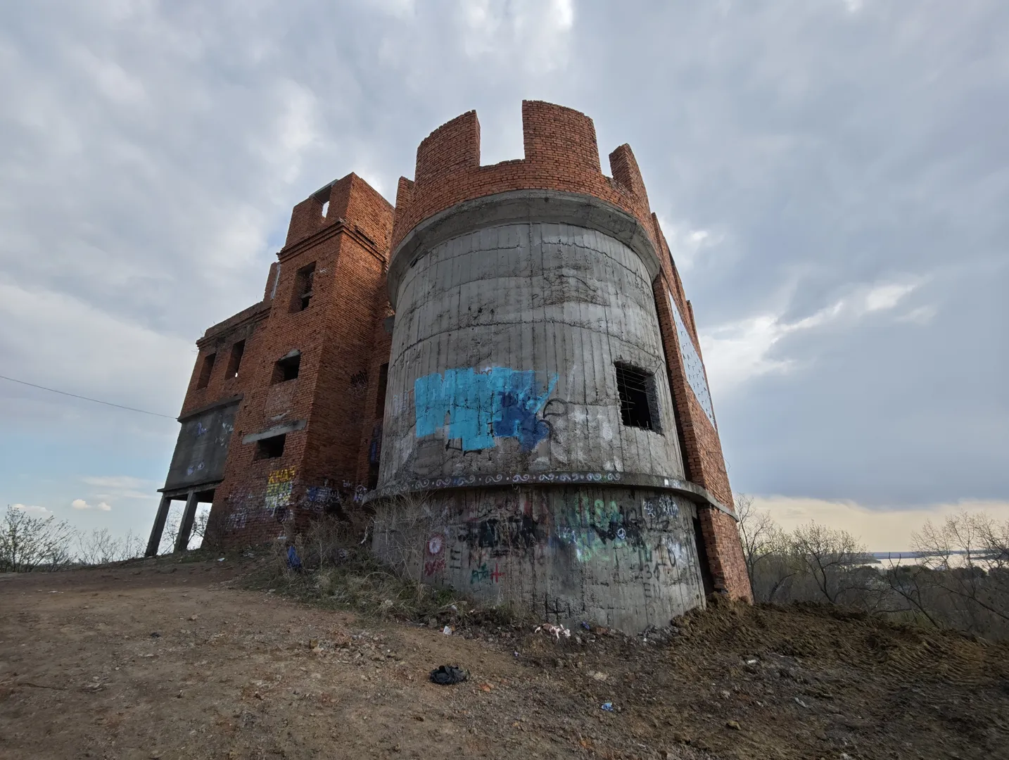 An ancient brick tower stands on elevated ground. The tower is covered with blue graffiti and has multiple windows. The structure is sturdy, surrounded by barren land and a small amount of vegetation.