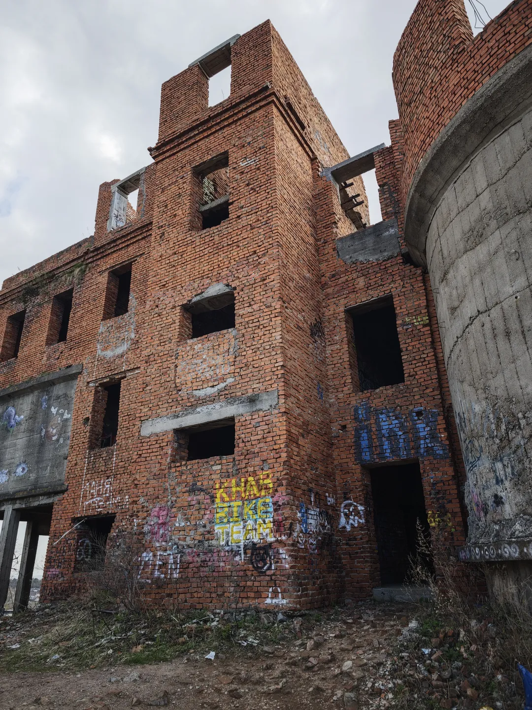 The ruins of a red brick building, with multiple window holes and graffiti on the walls. Next to the building is a gray concrete structure. The sky is overcast, with thick clouds.