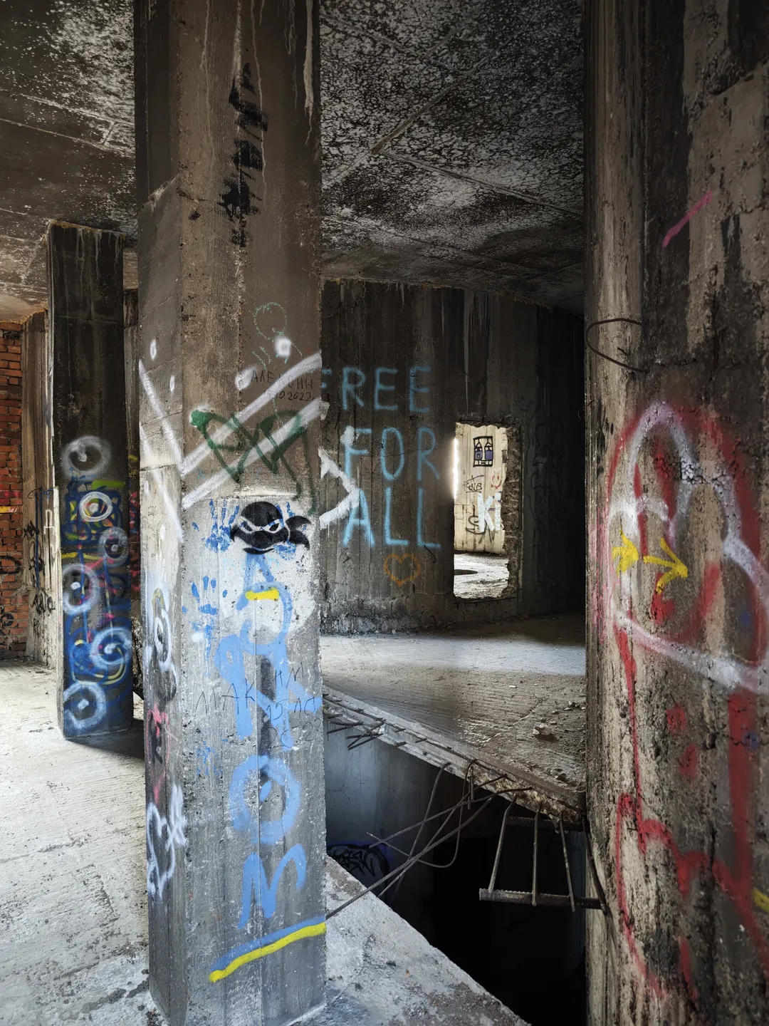 Inside the abandoned building, concrete columns are covered with colorful graffiti, with one column featuring the words “FREE FOR ALL”. The floor is damaged, and a window is visible in the distance.