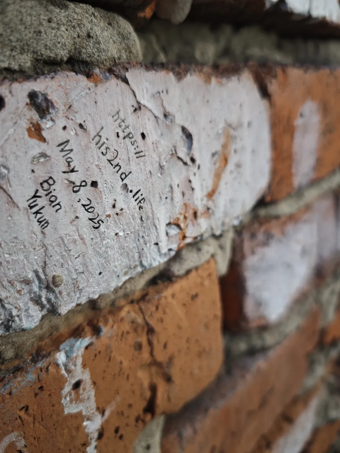 On a brick wall, the text “https://his2nd.life May 8, 2025 Bian Yukun” is engraved. The bricks are a mix of red and white. The cement parts of the wall have weathered and peeled off.