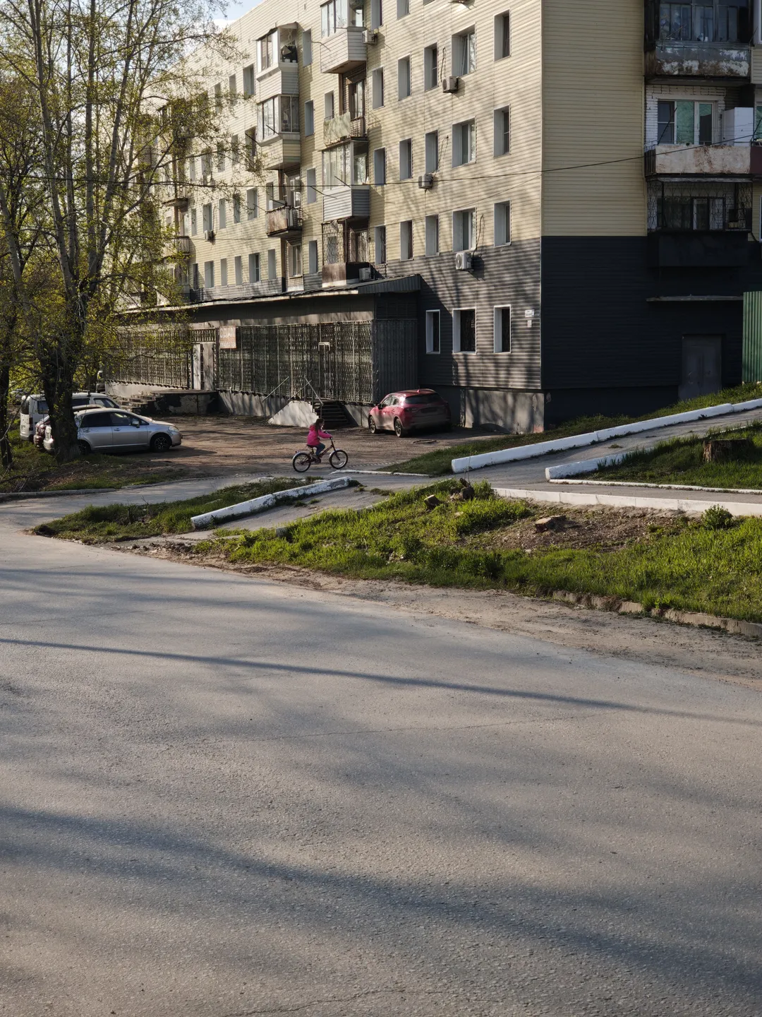A deserted road next to a multi-story residential building, with several cars parked in front. A child is riding a bicycle. Trees grow on one side of the road.