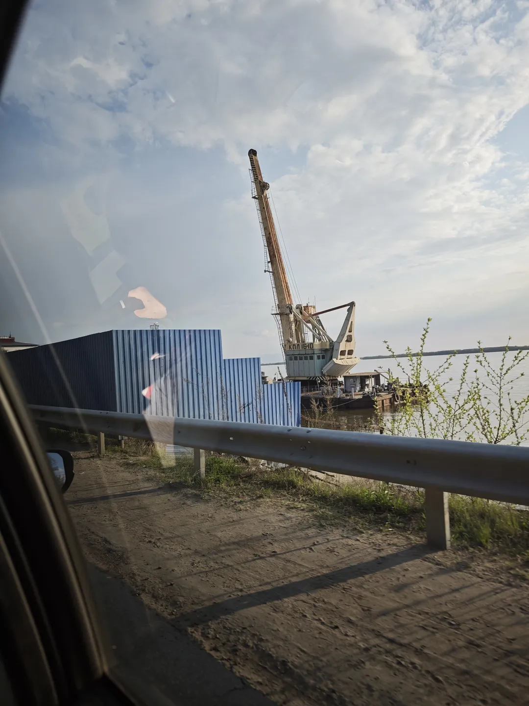 From inside a car, a view of a port can be seen, with a large crane and blue shipping containers. The sky is filled with white clouds, and the water is calm.