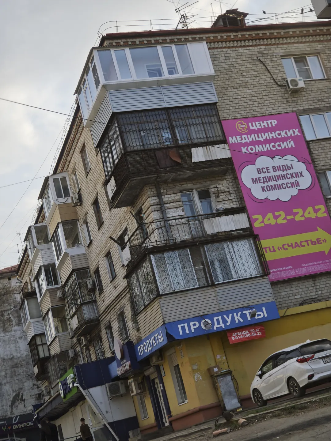 A multi-story building with a pink billboard on its exterior, featuring the words “ЦЕНТР МЕДИЦИНСКИХ КОМИССИЙ”. The ground floor shop is labeled “ПРОДУКТЫ”, and a white car is parked in front.