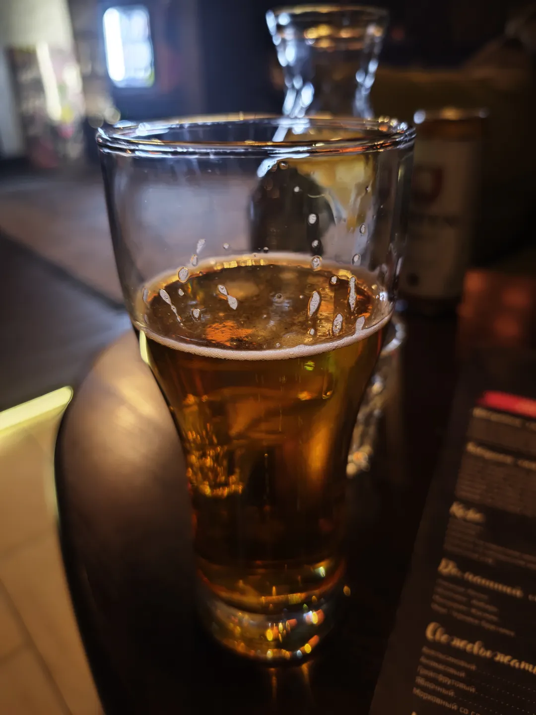 A glass of golden beer placed on a table, with foam on the surface of the beer. In the background, menus and other utensils can be seen. The glass reflects surrounding light.