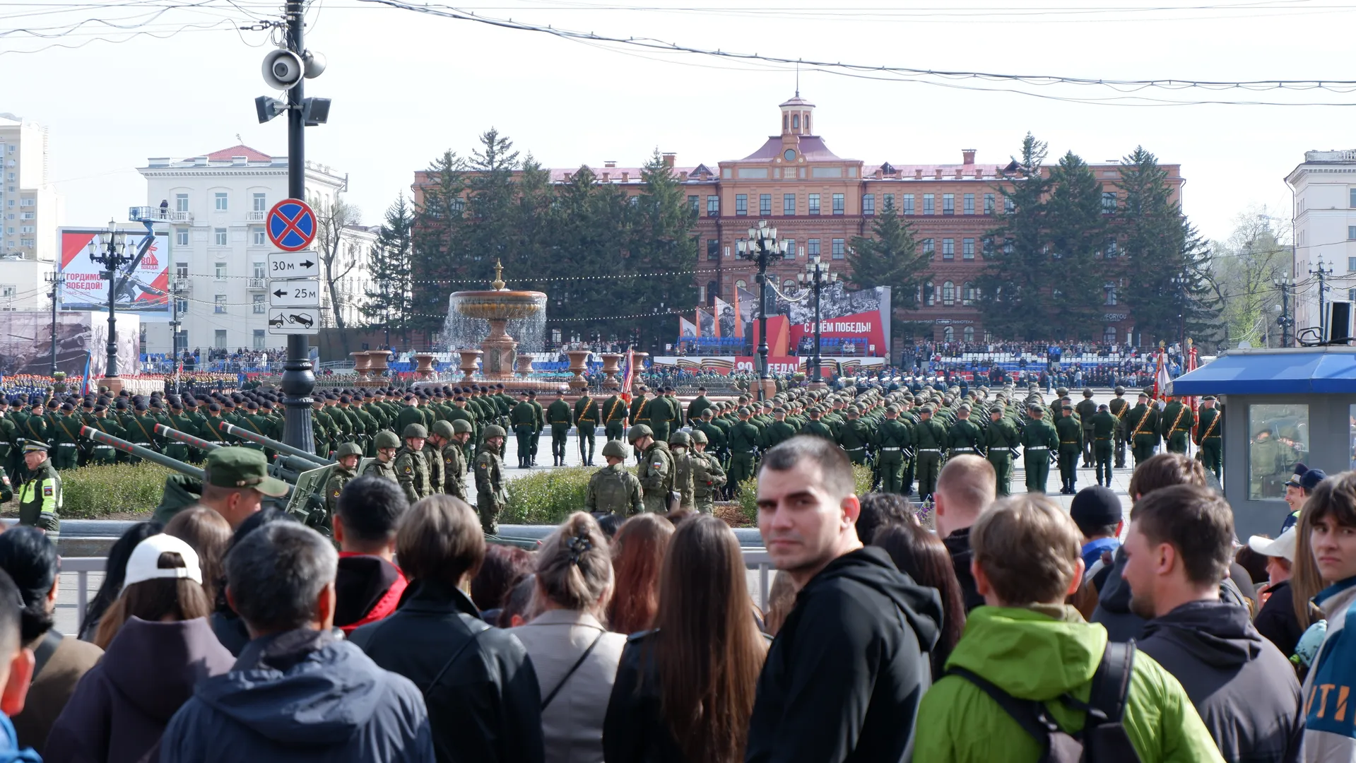 A large number of soldiers are lined up in the square, with a fountain and spectators in front. In the background, there are red buildings and green trees. Some spectators are wearing black coats, while others are wearing white hats.