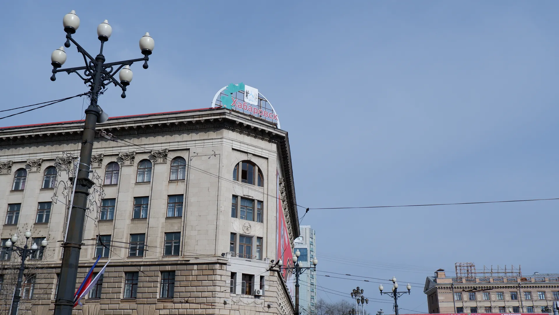 A corner of a multi-story building, with the word “Хабаровск” at the top. There are streetlights and power lines in front of the building. The sky is clear.