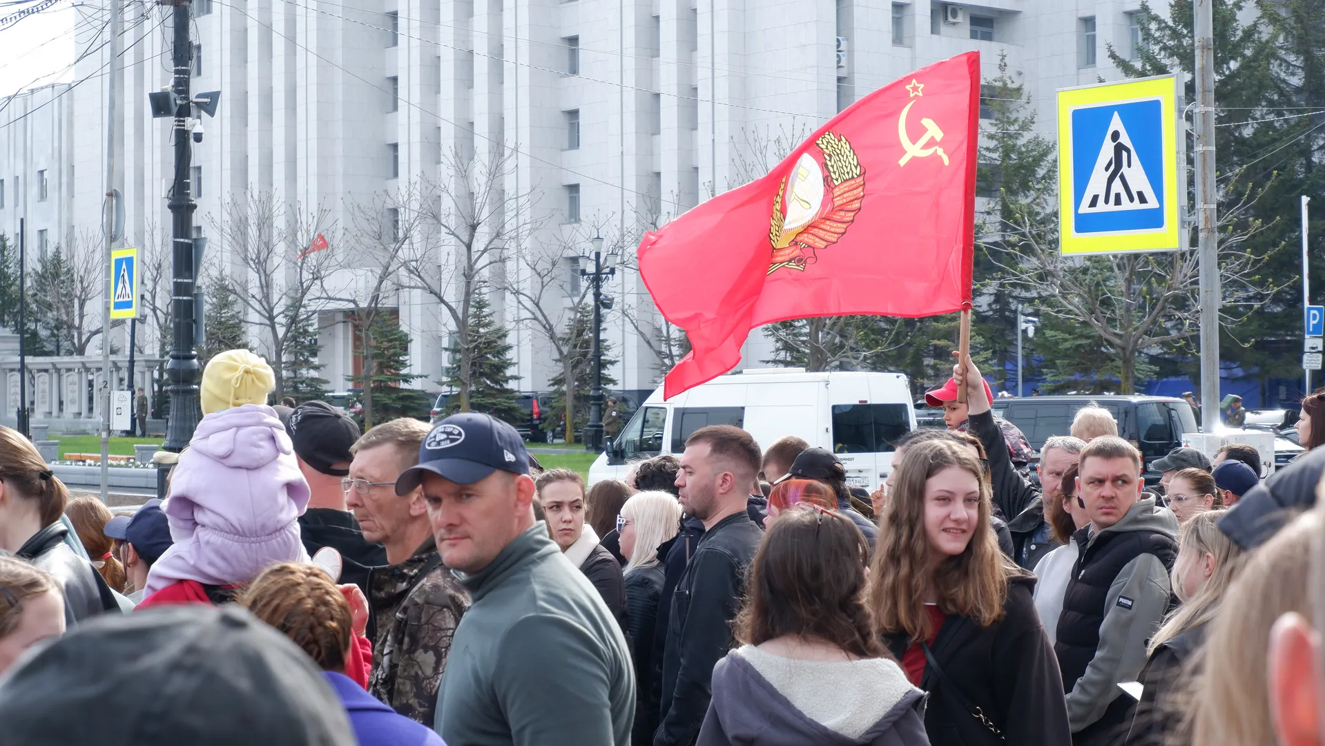 One person in the crowd is holding a red flag with the hammer and sickle symbol. In the background, there are tall buildings and bare trees. Pedestrians are wearing various coats.