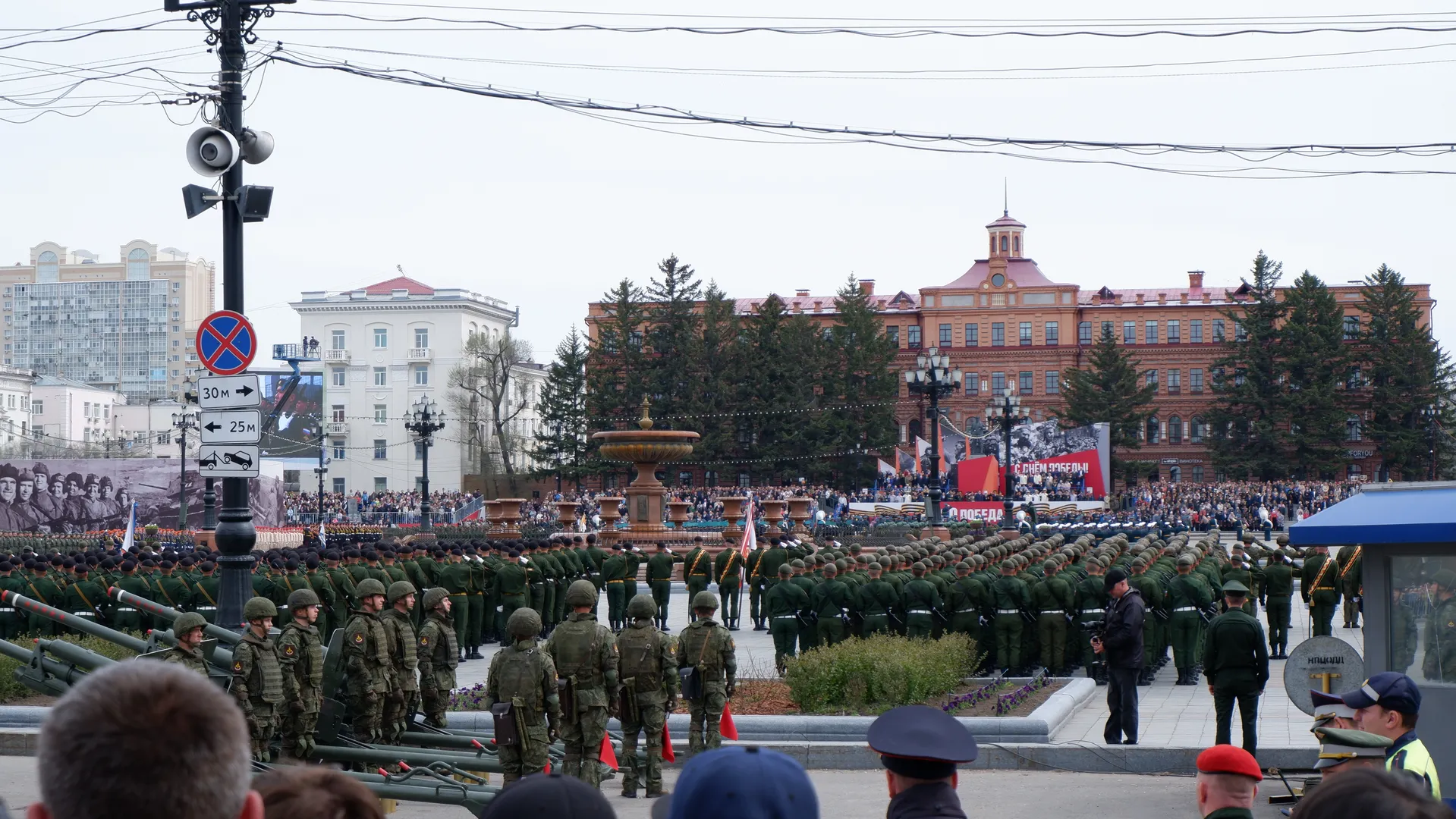 Soldiers are neatly arranged in the square, with large weapons and equipment in front. In the background, there are fountains and spectators. The buildings are red and white.