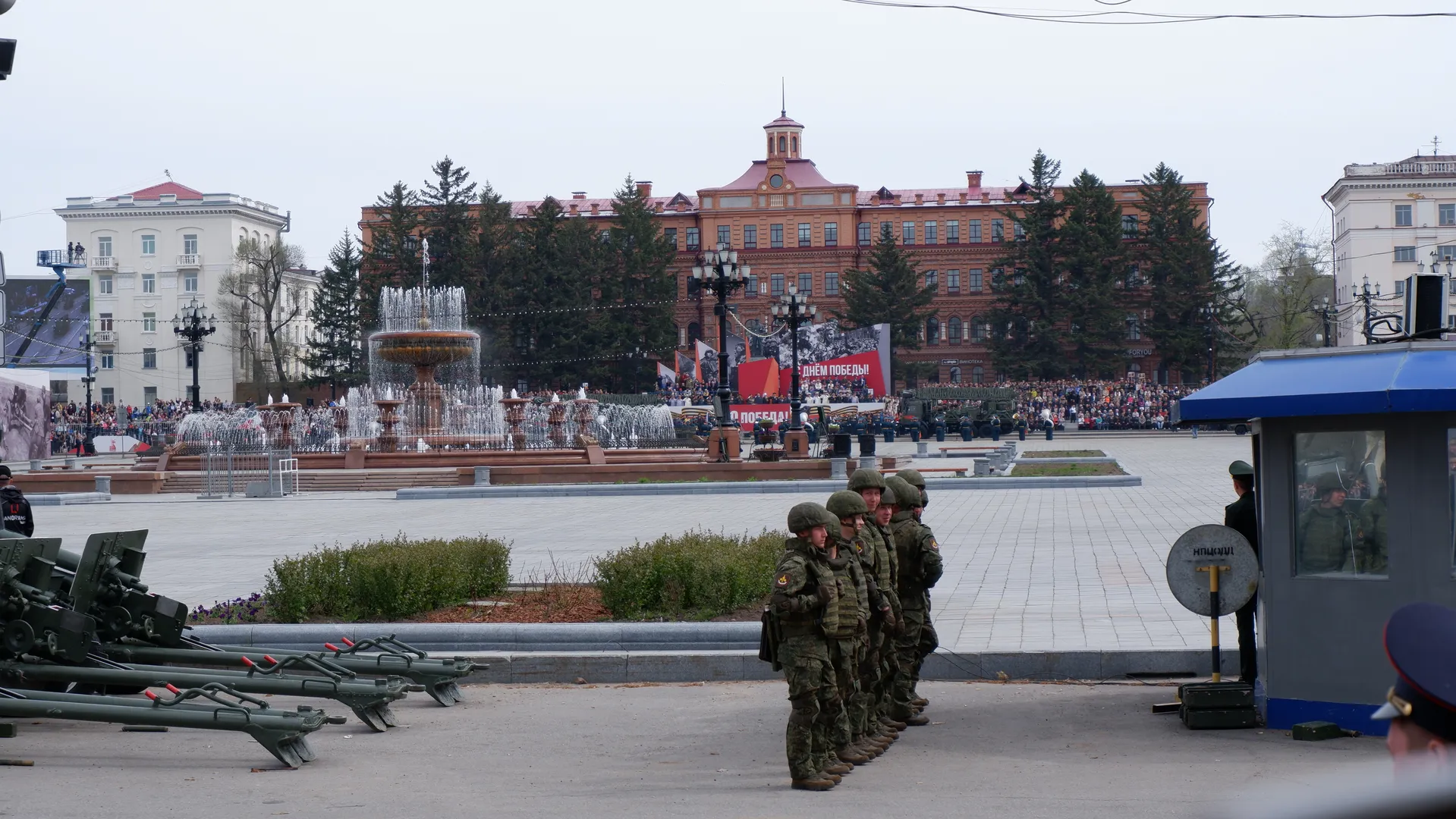 A fountain is located in the center of the square, with water splashing. Soldiers are lined up in front of the fountain, with spectators and red buildings in the background. A policeman is standing next to a kiosk.