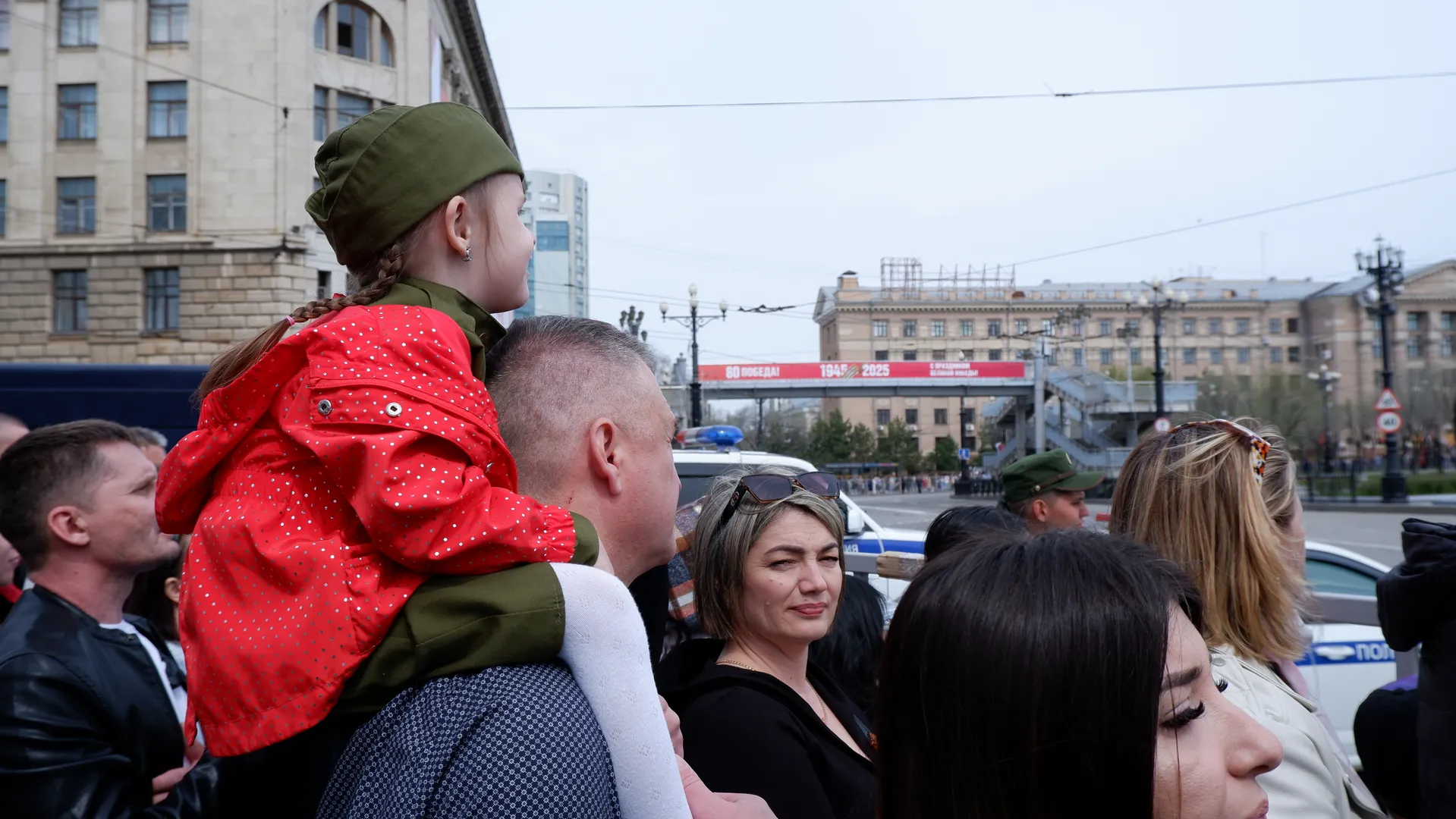 A child in a red coat is sitting on an adult’s shoulder, surrounded by a dense crowd. In the background, there are tall buildings and vehicles. A woman wearing sunglasses is smiling.
