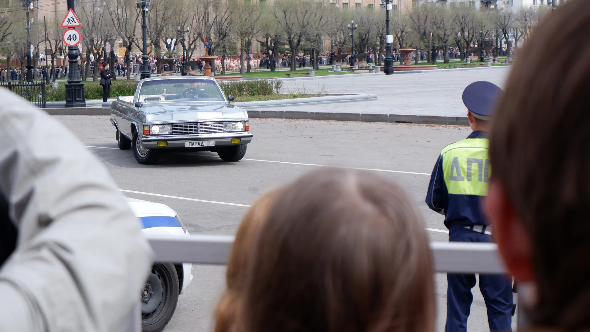 A convertible car is passing by with two people inside. There is a policeman directing traffic in front, and spectators are watching nearby. The background shows trees and buildings.