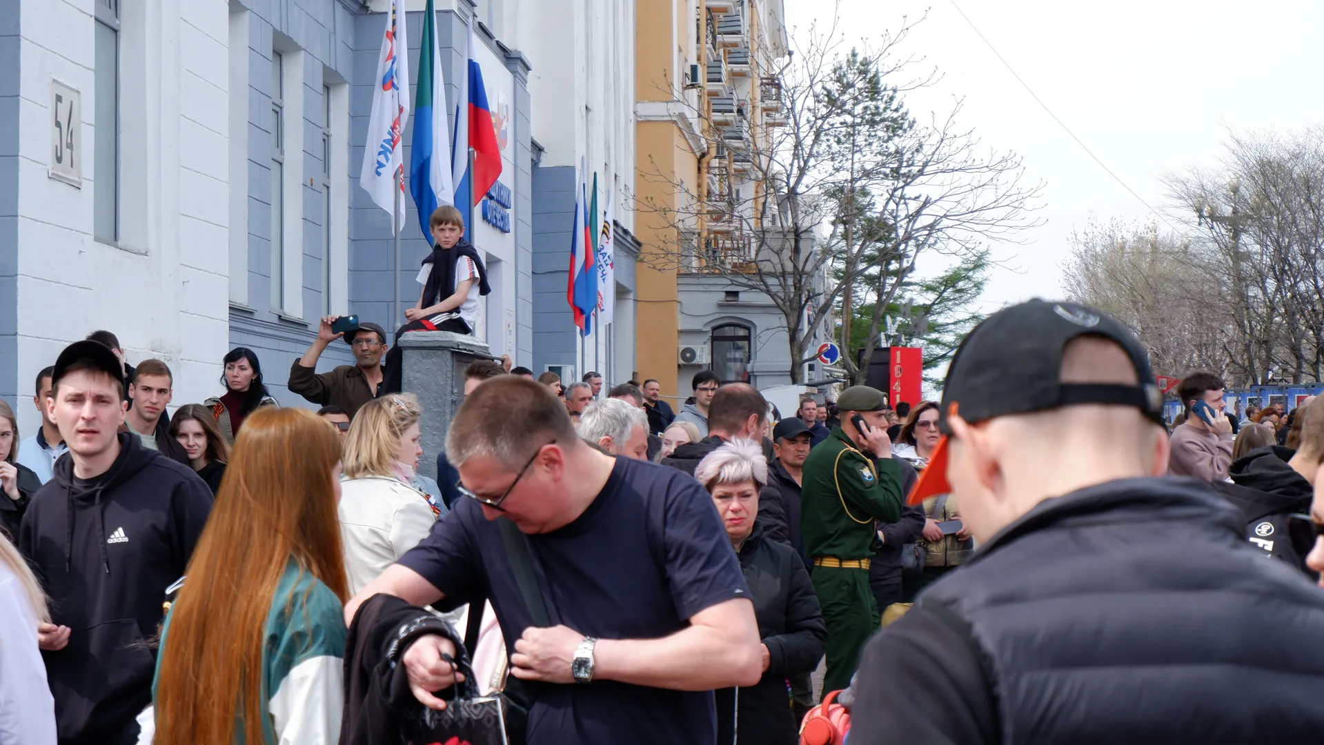 The street is crowded with people, one person is sitting in a corner taking pictures. In the background, there are flags and buildings. A man in military uniform is talking on the phone.
