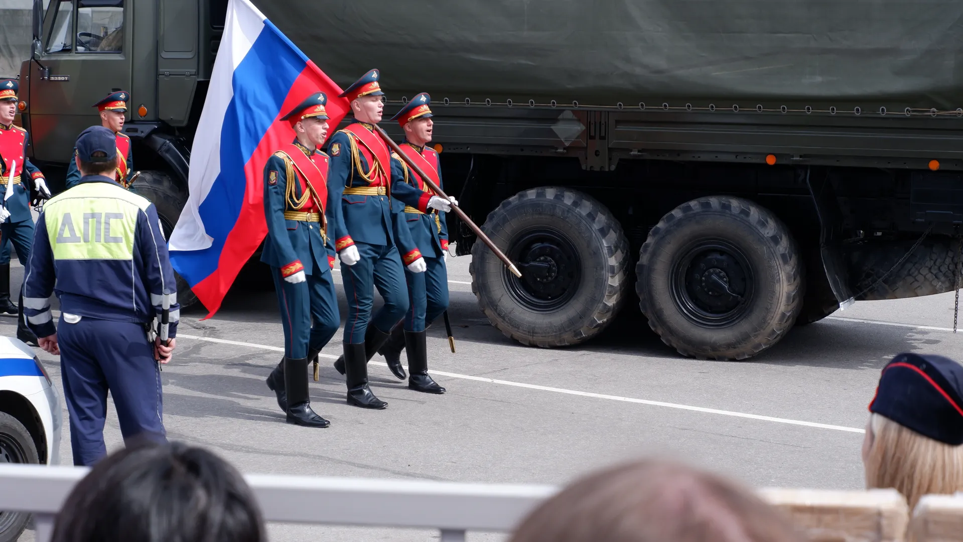 Several soldiers are marching, holding the Russian flag and wearing blue uniforms. Behind them is a large military truck. A policeman is standing by observing.