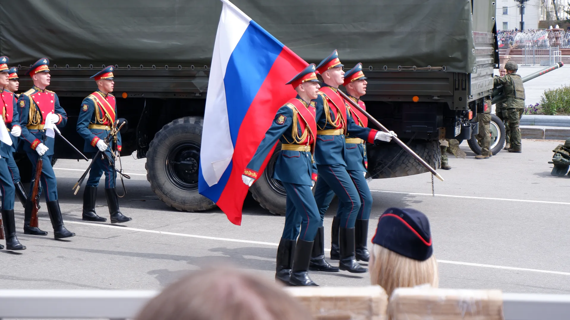 Several soldiers are marching, holding the Russian flag and wearing blue uniforms. Behind them is a large military truck. One soldier is adjusting equipment on the truck.