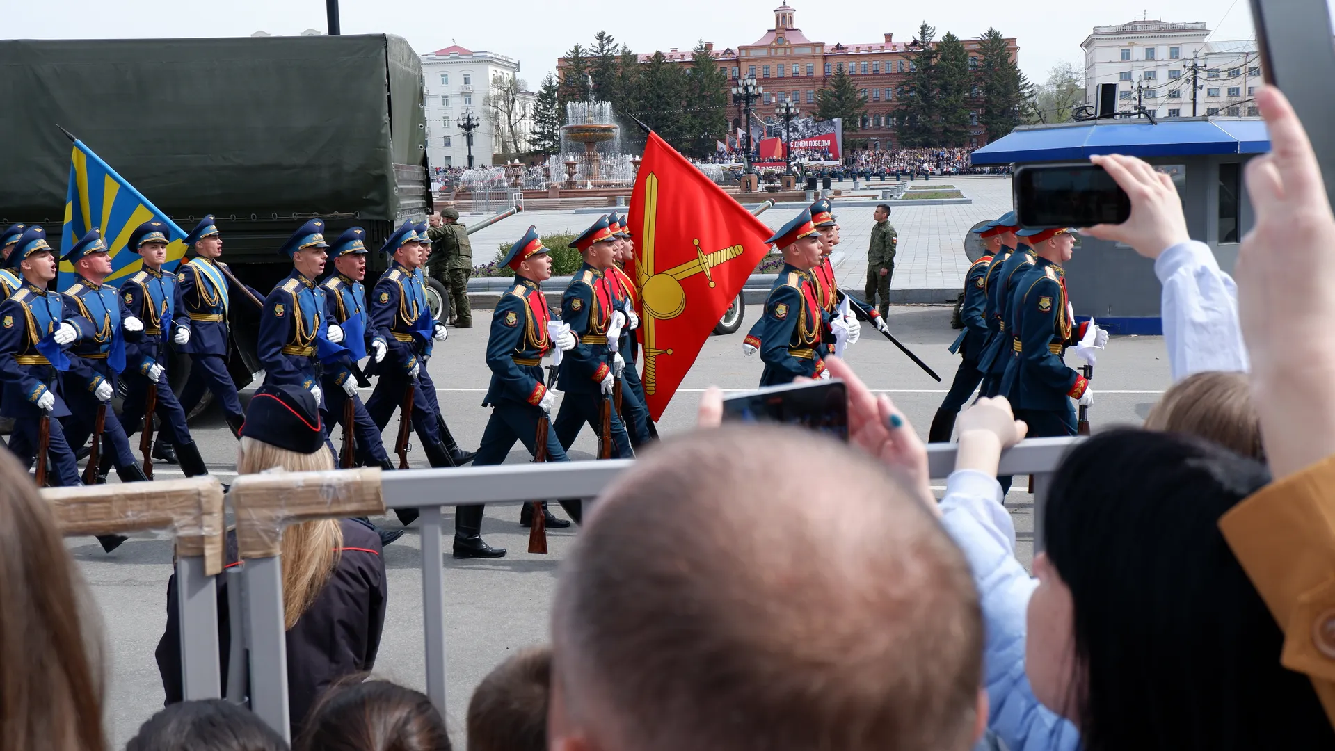 A group of soldiers in blue uniforms is marching, holding flags and rifles. In the background, there is a military truck and a fountain. Spectators are filming the scene behind the fence.