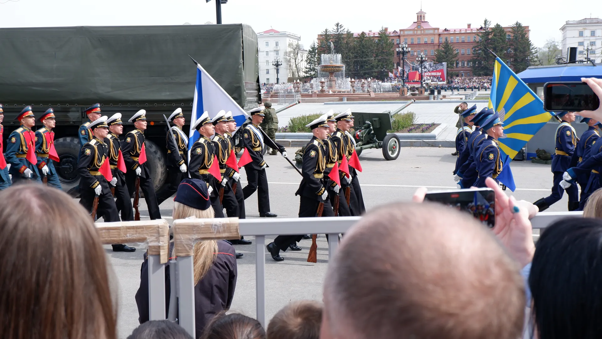 Two rows of soldiers are marching, each holding flags of different colors. The formation is neat and orderly. In the background, there are buildings and a fountain, with spectators filming in front. The soldiers’ uniforms have golden decorations.