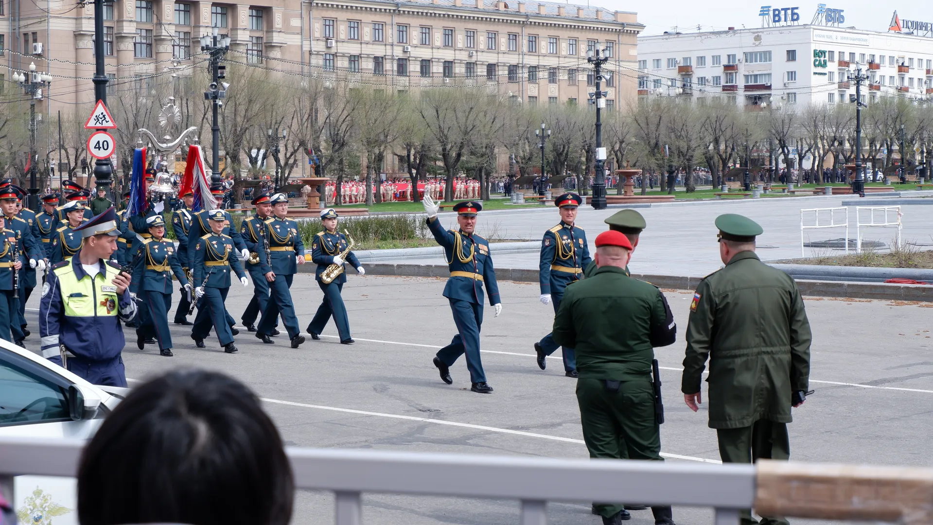 A military band is marching, with members wearing dark uniforms and hats. In front, there are two individuals in green military uniforms. The background shows city buildings and trees.