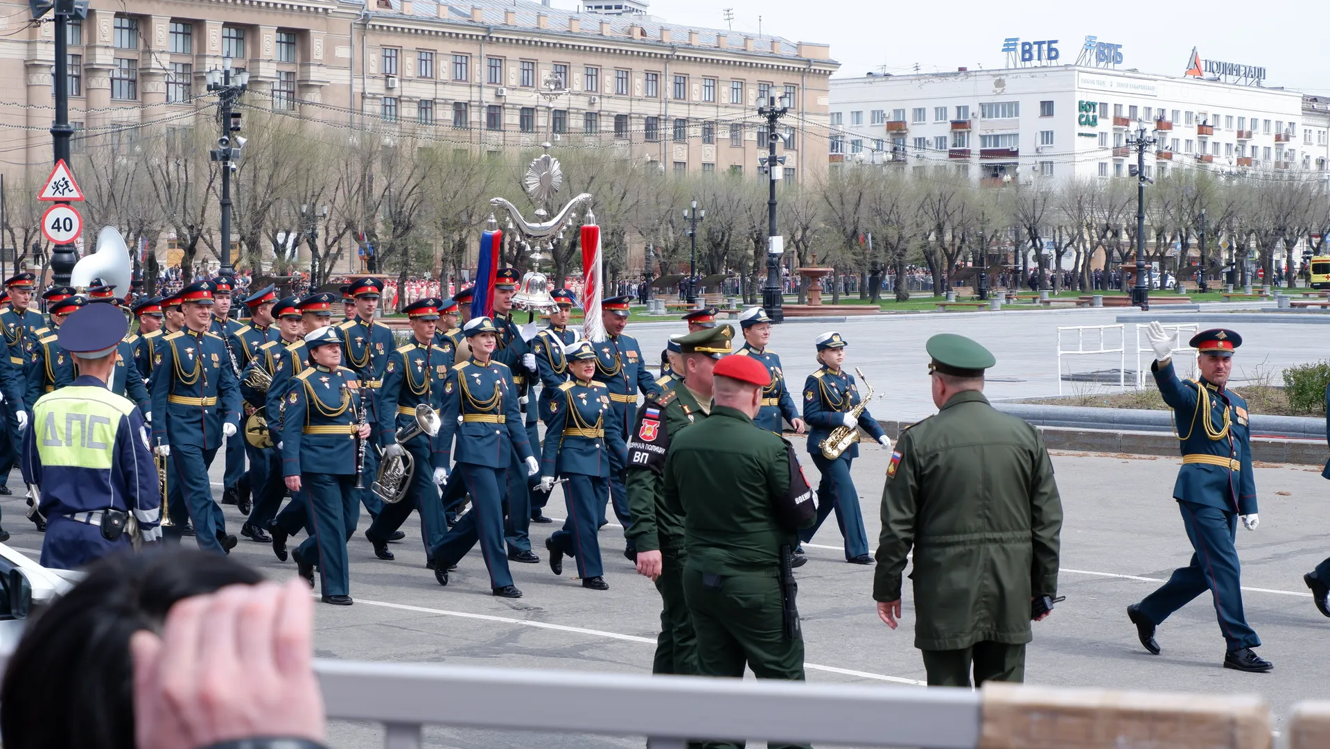 The military band continues marching, with members holding instruments in sync. The individuals in green military uniforms ahead have their backs to the camera. The background clearly shows buildings and trees.